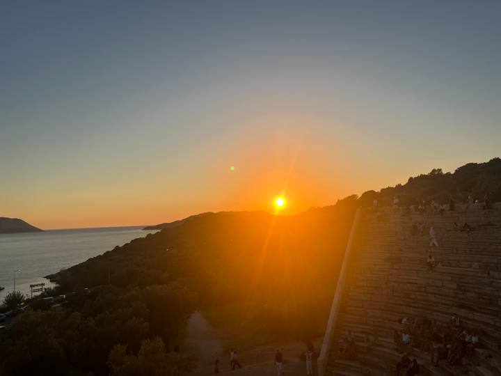 Ancient hillside amphitheatre filled with visitors watching the sun set over the sea.