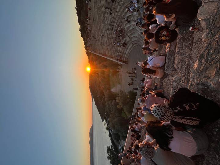 Large crowd seated in an ancient theatre at sunset with the sun dipping toward the sea.