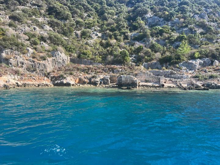 Rocky coastline with clear blue water and sparse ancient stone remains at the shore.