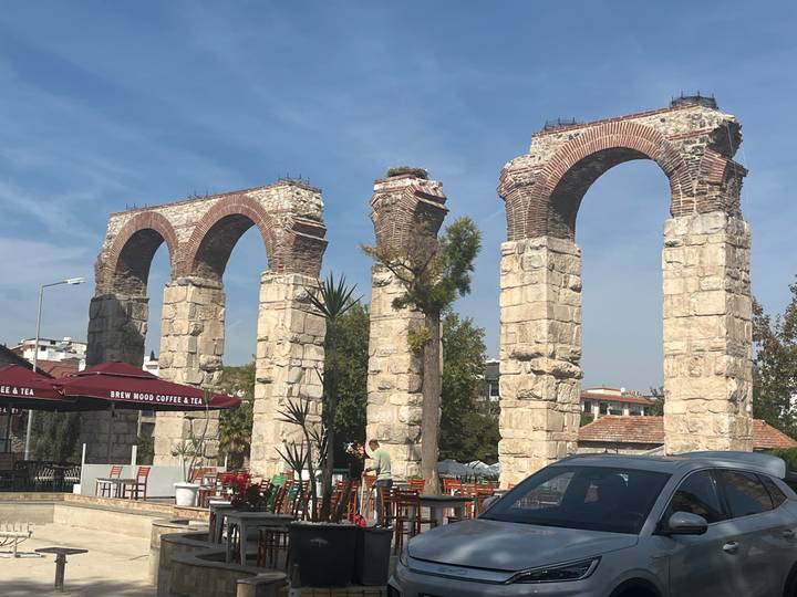 Three surviving arches of an ancient aqueduct beside a modern café terrace and parked car.