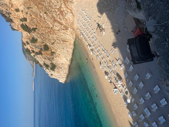 Idyllic sandy cove with turquoise water, lined with neat rows of white sun loungers beneath steep cliffs.