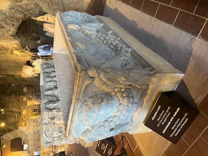 Ancient sarcophagus with detailed relief carvings displayed in a museum with visitors nearby.