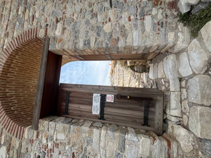 Wooden gate within a stone wall opening to ancient ruins, with a small notice sign attached.