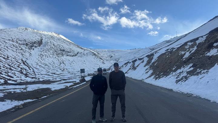 Two men stand on a deserted mountain road flanked by snowy slopes and a blue sky.