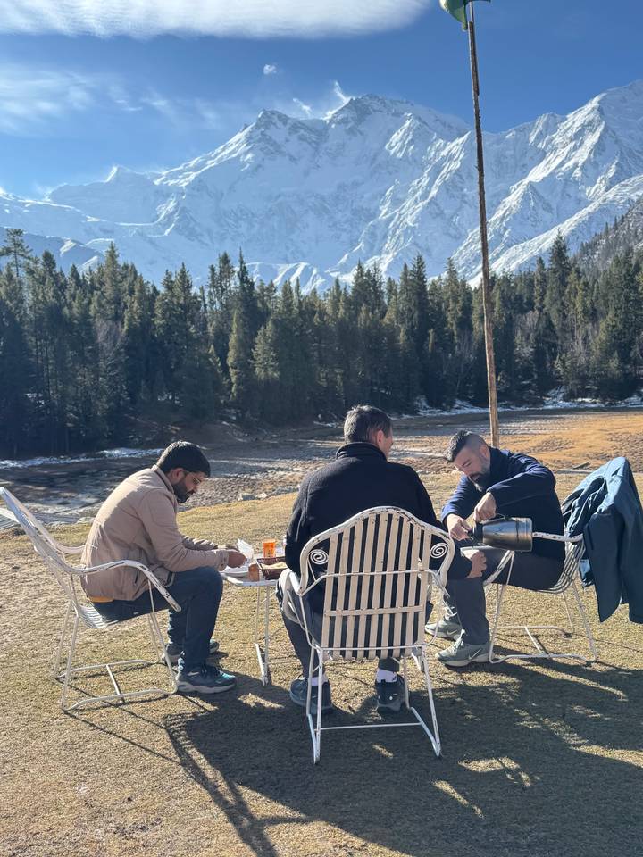 Three men sit outdoors enjoying tea with snow-capped peaks and pine trees in the background.