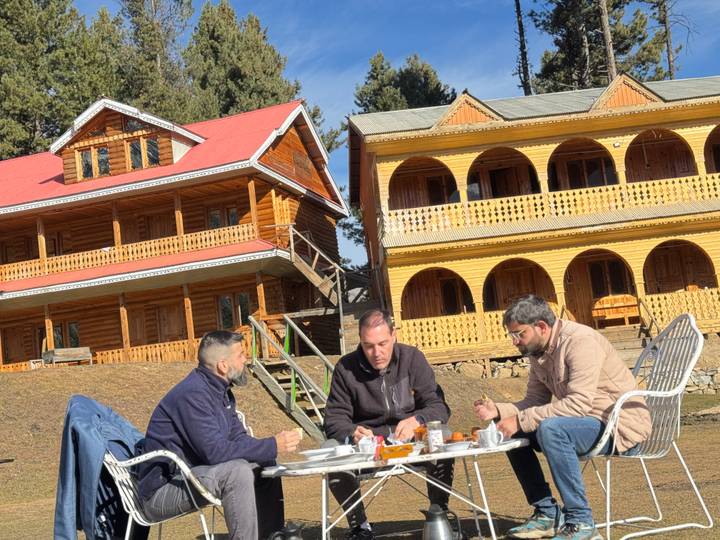 Three men have breakfast outside wooden mountain lodges in bright sunshine.