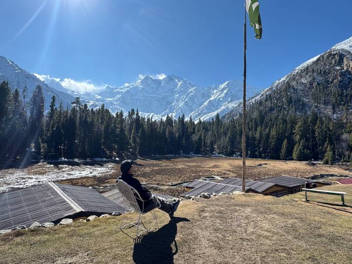 A lone traveler relaxes on a chair facing snow-covered peaks and dense pine forest at Fairy Meadows.