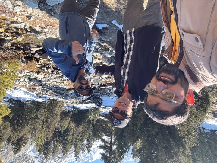 Three men pose for a selfie near a rocky stream surrounded by evergreens and patches of snow.
