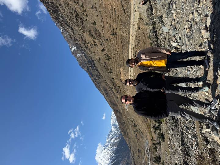 Three men stand on rocky terrain with towering, snow-tipped mountains rising behind them.