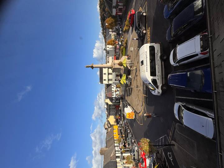 Street-level view of a small Irish town square with monument, market stalls and parked vehicles under blue sky.