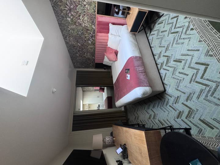 Modern hotel room with a double bed topped with pink accents, skylight above, desk and mirror at the far wall.