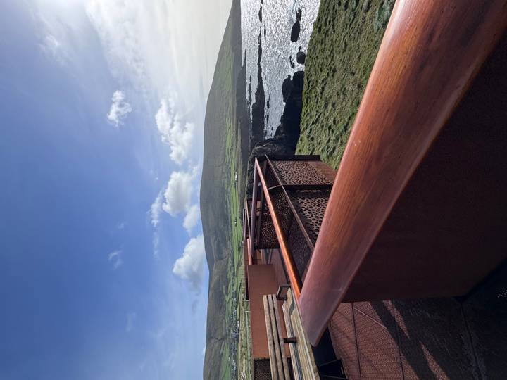 Coastal viewpoint deck overlooking rugged cliffs, crashing waves and green hills in bright sunlight.