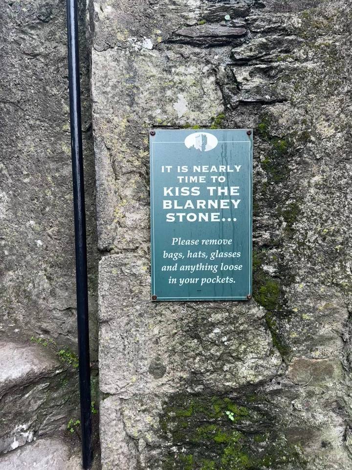 Green sign on stone wall advising visitors about kissing the Blarney Stone.
