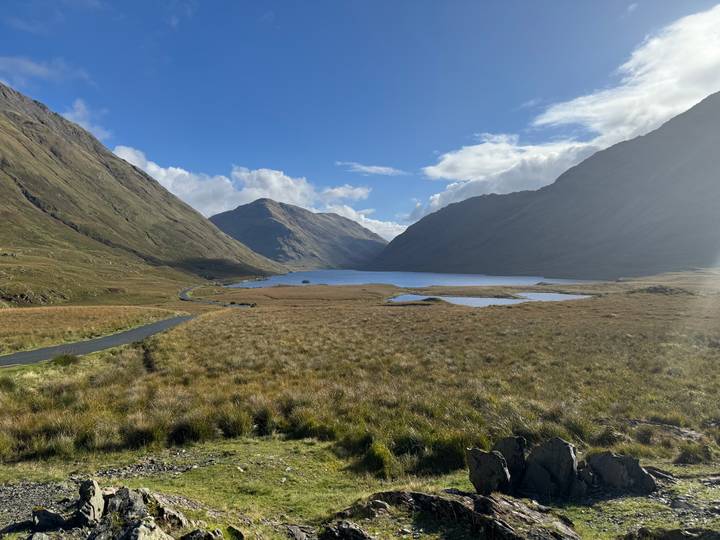 Expansive highland valley with winding road, mountain walls and a shimmering lake under blue sky.