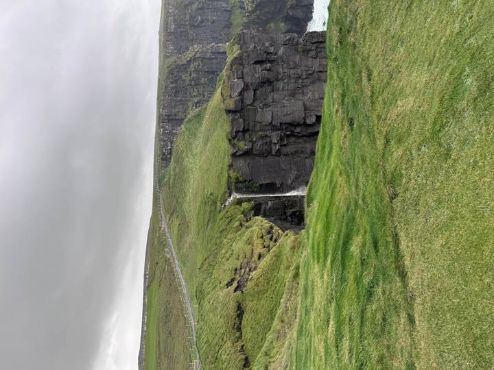 Green waterfall plunges over dramatic dark cliffs topped with windswept grass under gray sky.