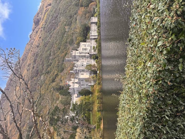 Grand Kylemore Abbey reflected in a calm lake, backed by forested slopes in autumn tones.