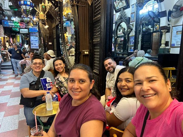 Friends relax with drinks inside a bustling traditional bazaar café lined with ornate woodwork.