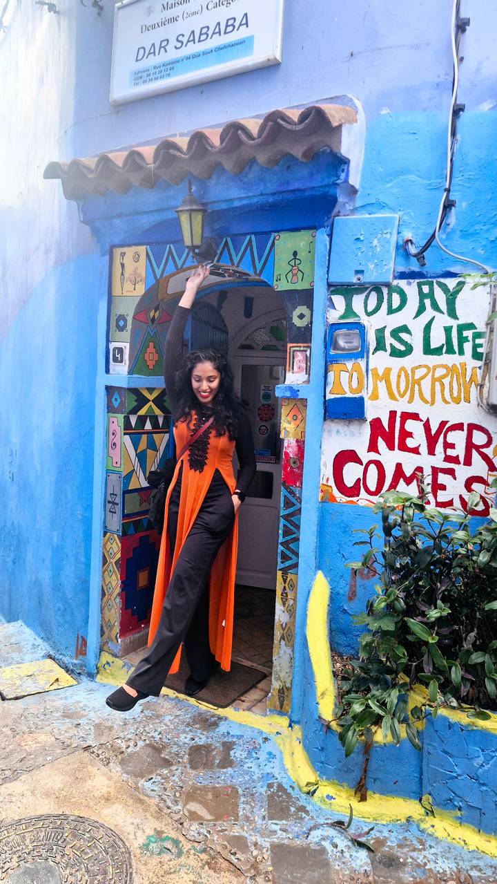 Smiling woman in orange dress stepping out of a vividly painted blue doorway covered with colorful Berber patterns and slogans.