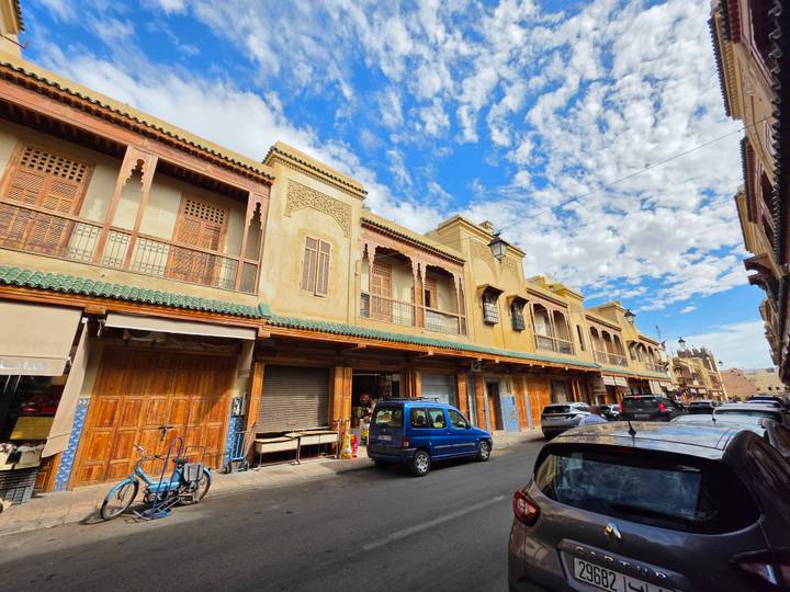 Traditional souk street lined with tan buildings, wooden shutters and green-tiled roofs under a bright blue sky; locals and vehicles below.