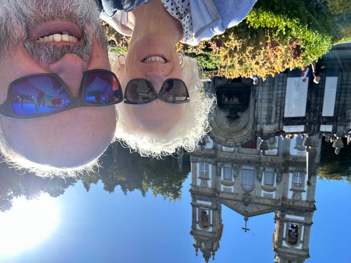Older couple taking a sunny selfie in front of the ornate twin-towered sanctuary building and formal gardens