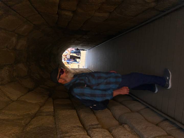 Older male traveller walking through a narrow stone tunnel toward daylight and a waiting group