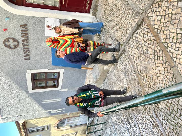Street scene with travellers posing next to a person in a colourful traditional costume under a Lusitania sign