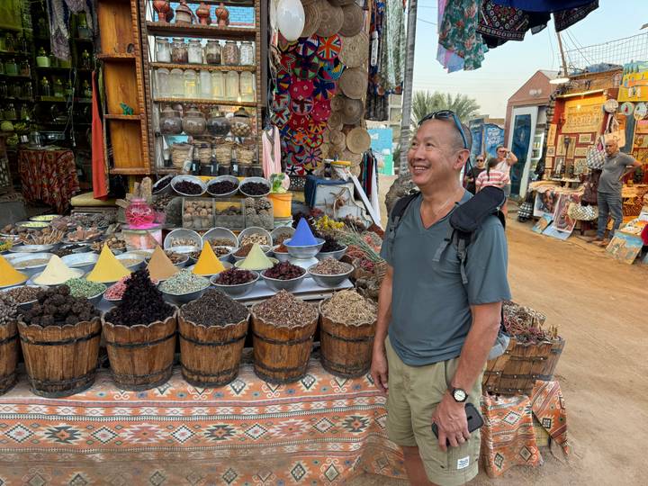 Smiling tourist stands beside colorful barrels and pyramid piles of spices at an Egyptian street market stall.