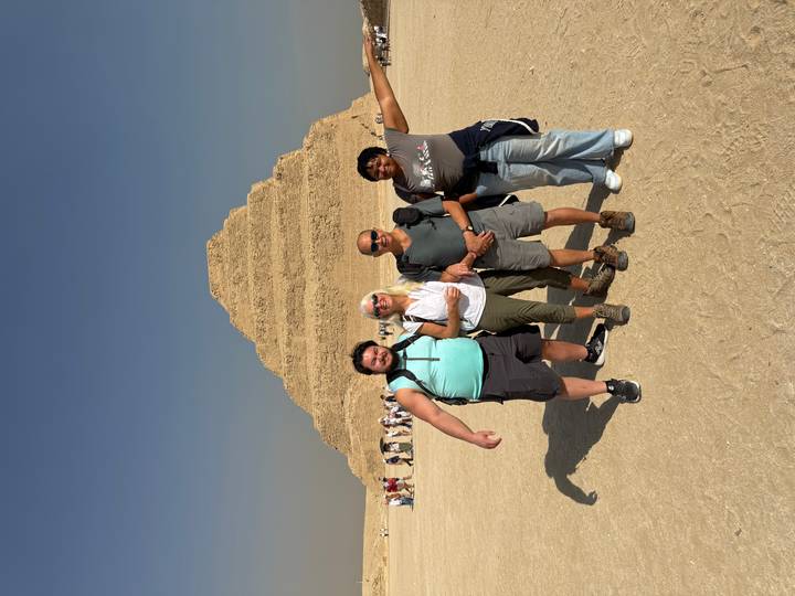 Four travelers pose arm-in-arm in front of the Step Pyramid of Saqqara on a bright desert day.