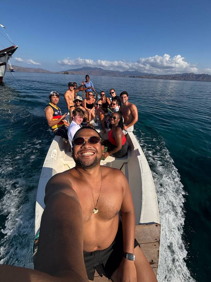 Large group of friends smile on a small speedboat cutting through clear blue water under bright sunlight.