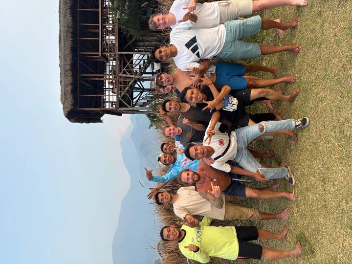 Travelers and local guides strike joyful poses on a grassy hillside with wooden stilt house and hazy volcanoes behind.