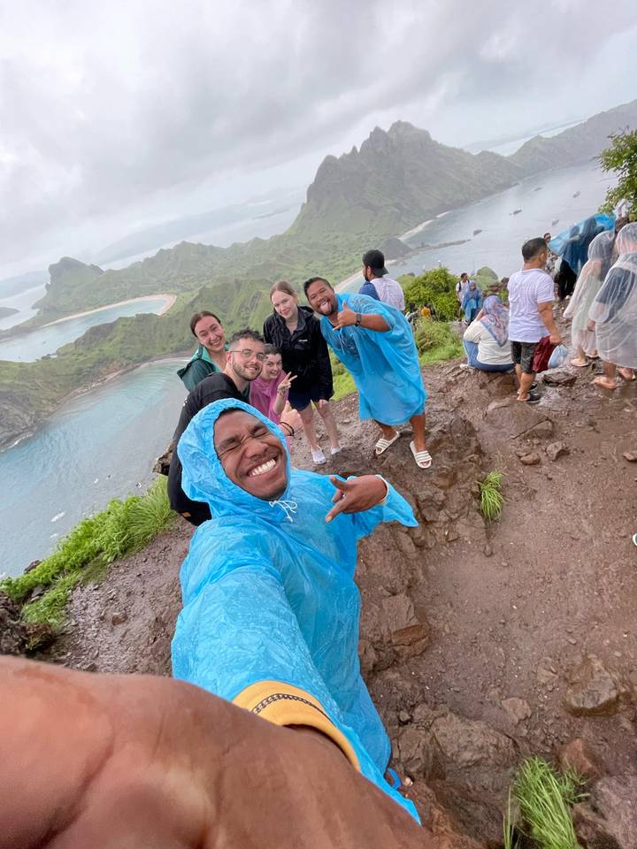 Wet but smiling hikers in blue ponchos posing on a coastal viewpoint overlooking bays and islands.
