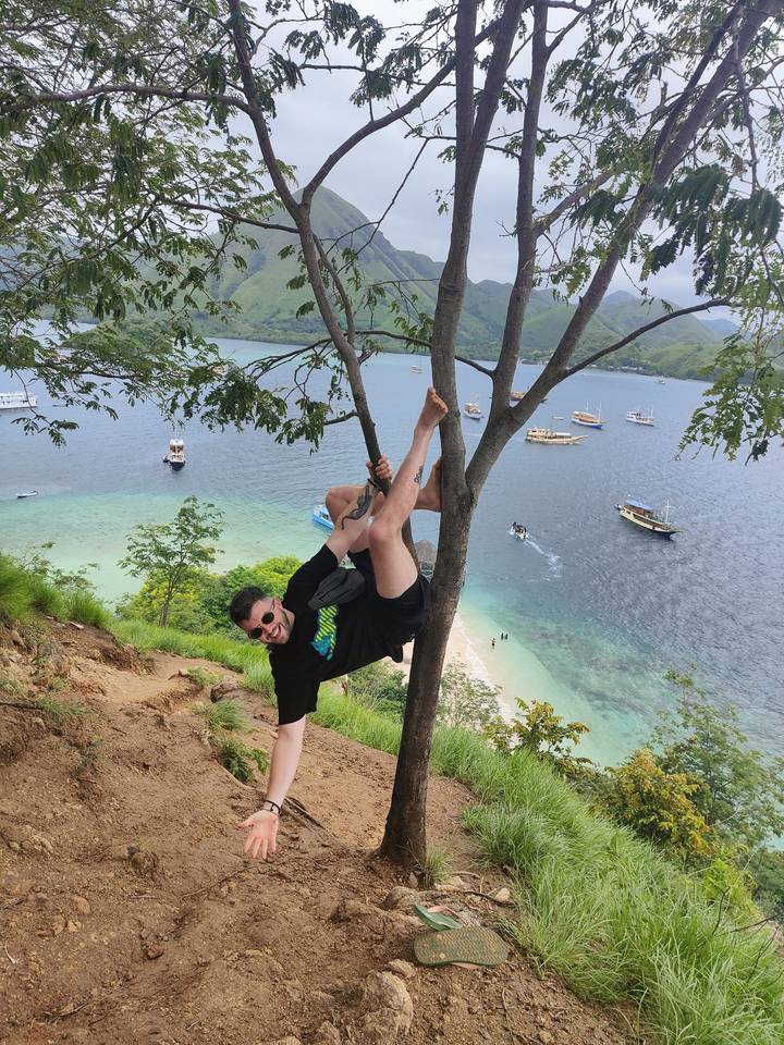 Traveler playfully hanging from a tree on a hill overlooking turquoise bays filled with boats.