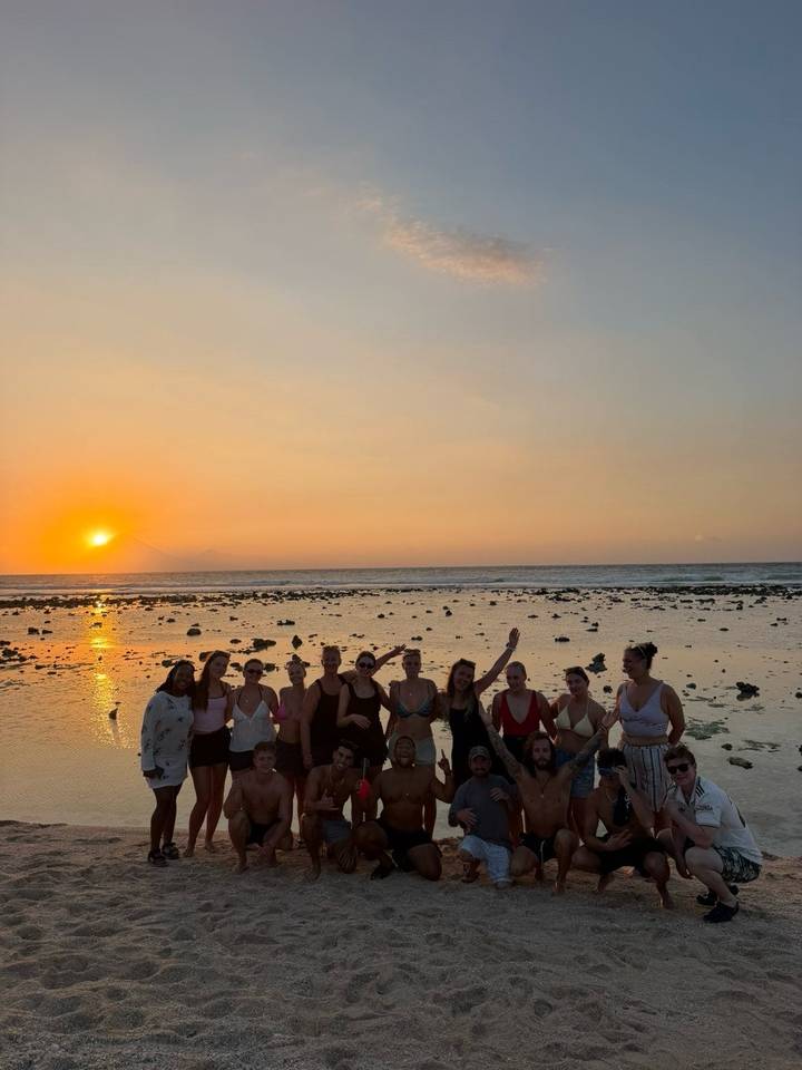 A line of young travelers posing on a rocky beach at sunset with the orange sun dropping over the sea horizon.