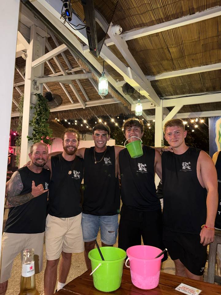 Five friends in matching black singlets smile at a lively bar decorated with string lights.