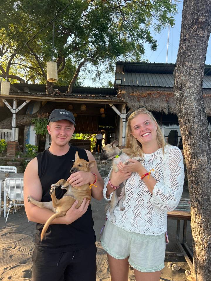Smiling couple seated outdoors cuddle two small rescue puppies in front of a rustic café.