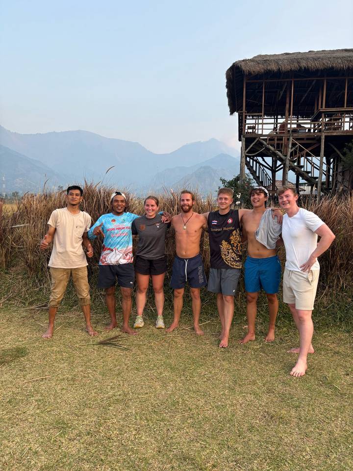 Seven friends stand arm-in-arm in tall grass with hazy mountains rising behind them.