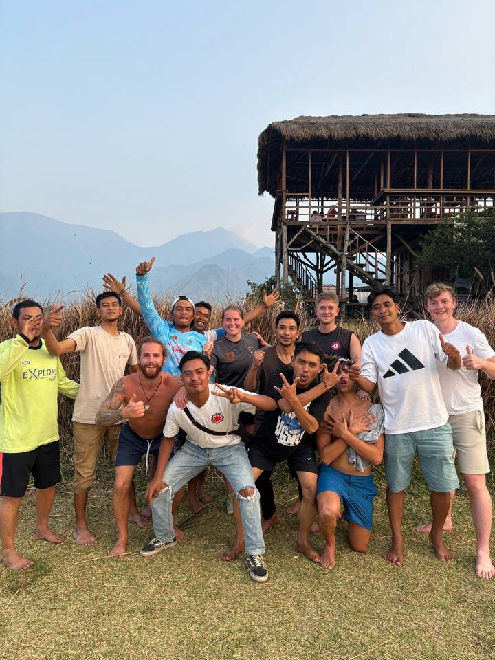 Tourists and local guides gesture playfully together beside a stilted wooden building near fields.