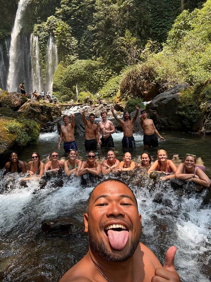 Group cools off in a rocky stream below a waterfall, leaning on the ledge and cheering to the camera.