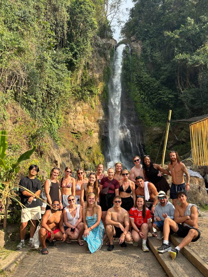 Travel group smiles at the base of a tall tropical waterfall surrounded by lush cliffs.