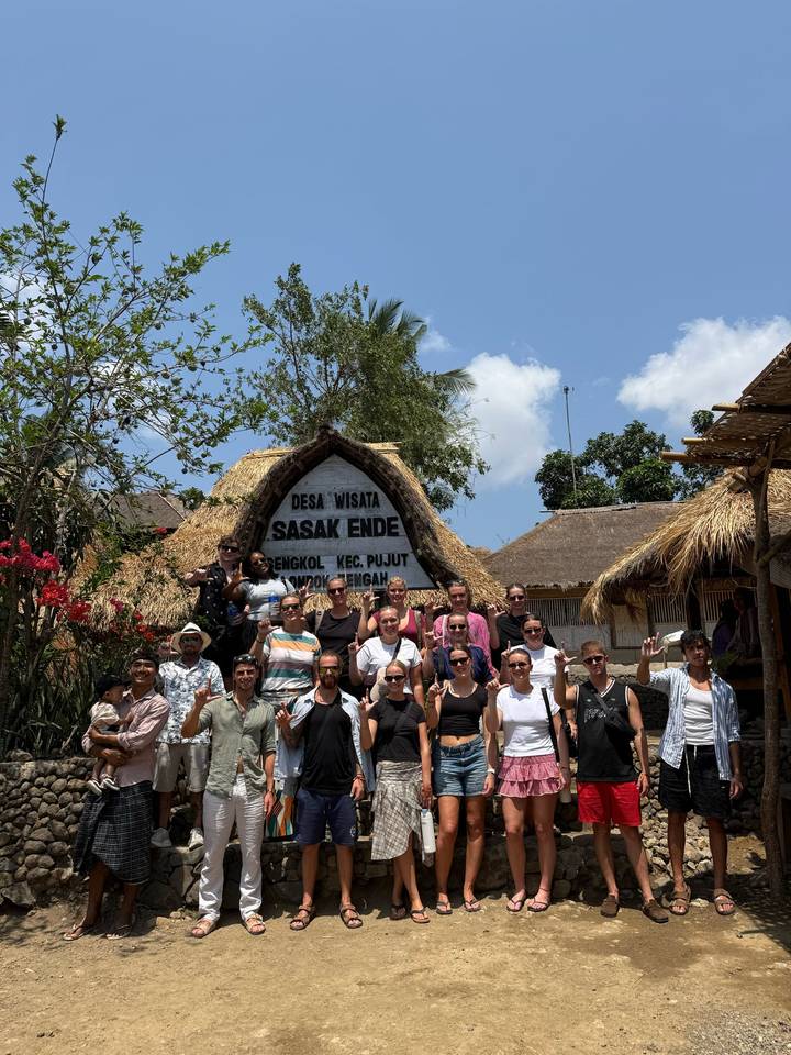 Large tour group poses under a thatched gate reading 'Desa Wisata Sasak Ende' in bright sunshine.