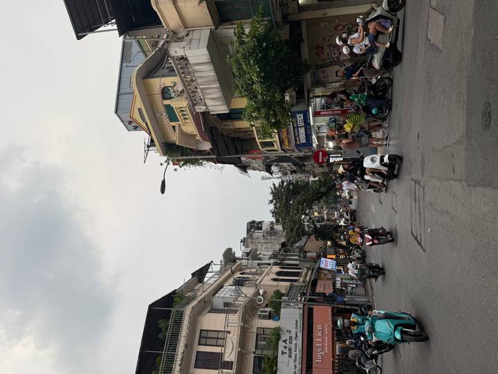 Bustling Vietnamese street lined with narrow buildings, scooters and pedestrians beneath an overcast sky.