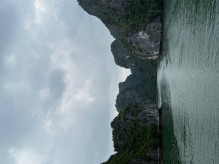 Narrow channel of emerald water flanked by towering forested karst cliffs beneath a cloudy sky.