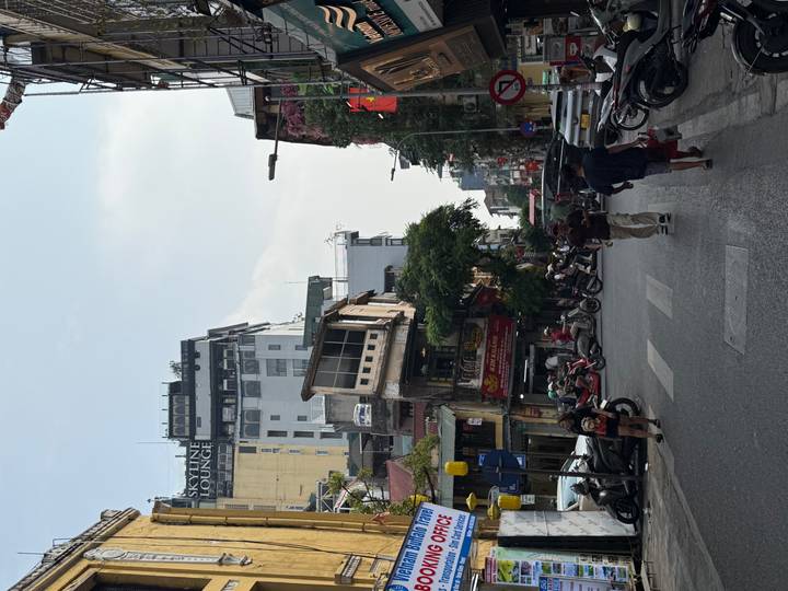 Busy Hanoi street scene with pedestrians, motorbikes, and tall shopfronts under a pale sky.