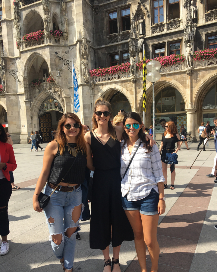 Trois femmes posant devant un bâtiment historique avec une enseigne munichoise.