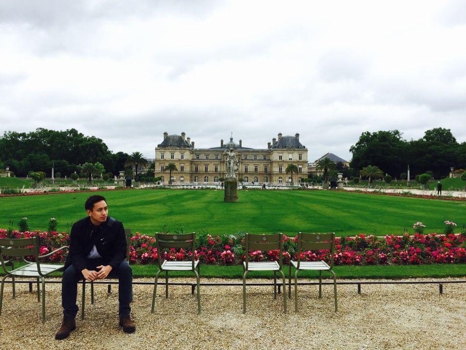 A person sitting on a chair in a manicured garden with a historic building in the background.