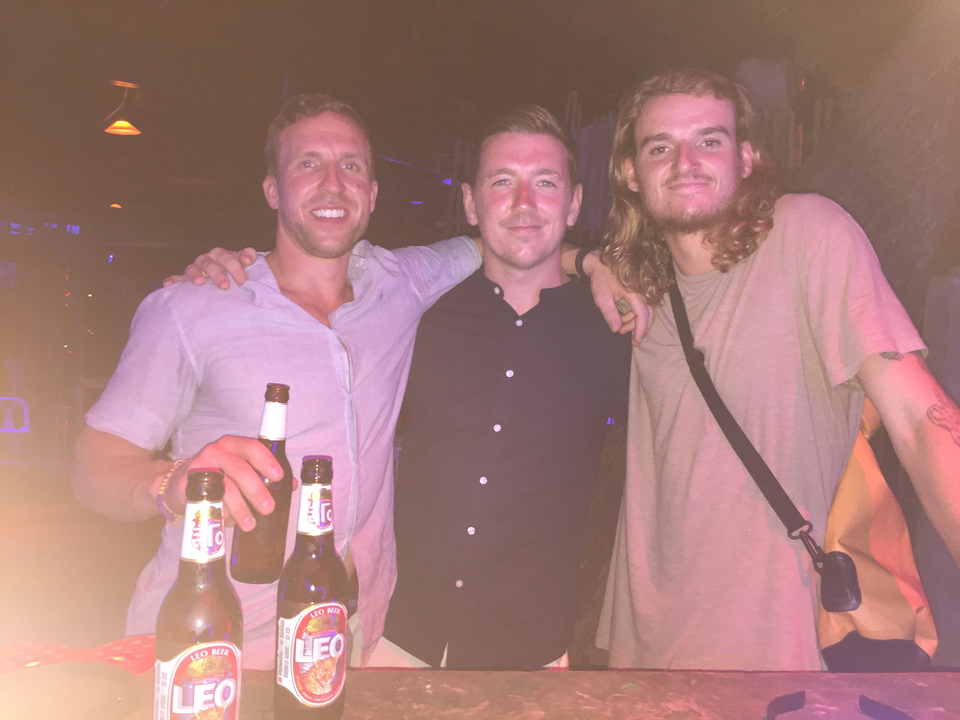 Three friends standing together while holding beer bottles in a lit room.