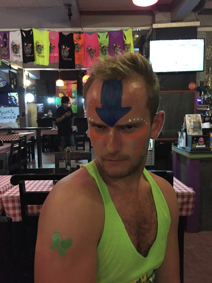 Young man with blue arrow face paint and neon accents posing inside a casual bar.