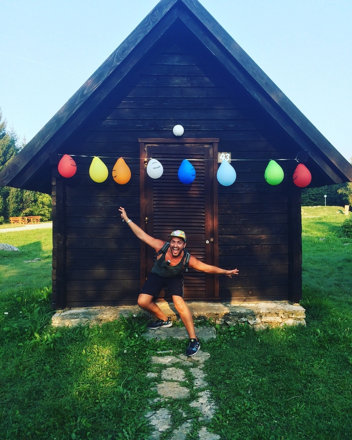 Personne posant avec enthousiasme devant un bâtiment en bois avec des ballons colorés.