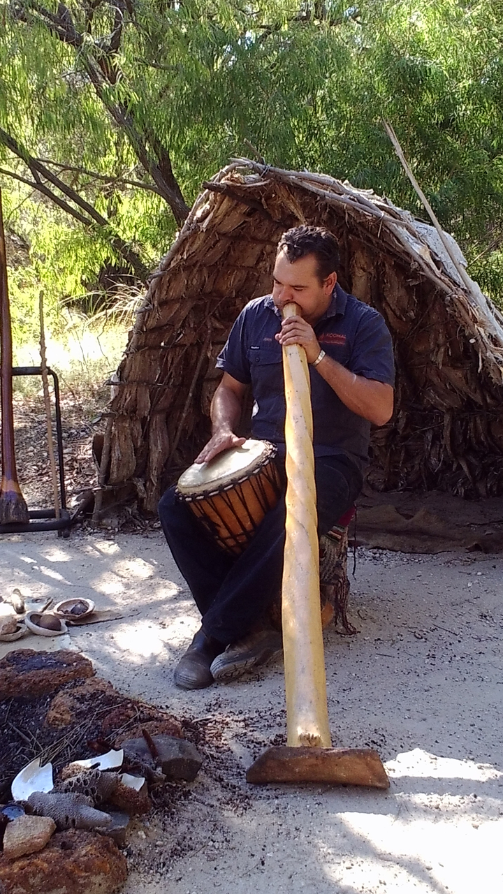 Une personne jouant du tambour et tenant un didgeridoo à côté d'une hutte tressée.