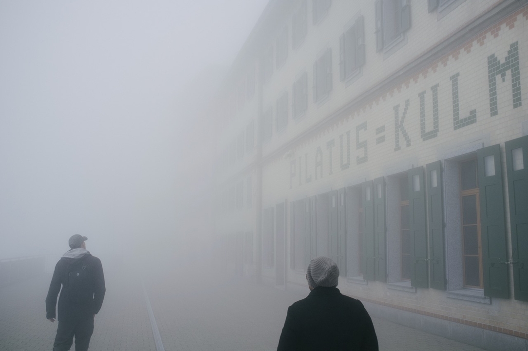 Personnes marchant dans une zone brumeuse, "Pilatus Kulm" écrit sur un bâtiment.